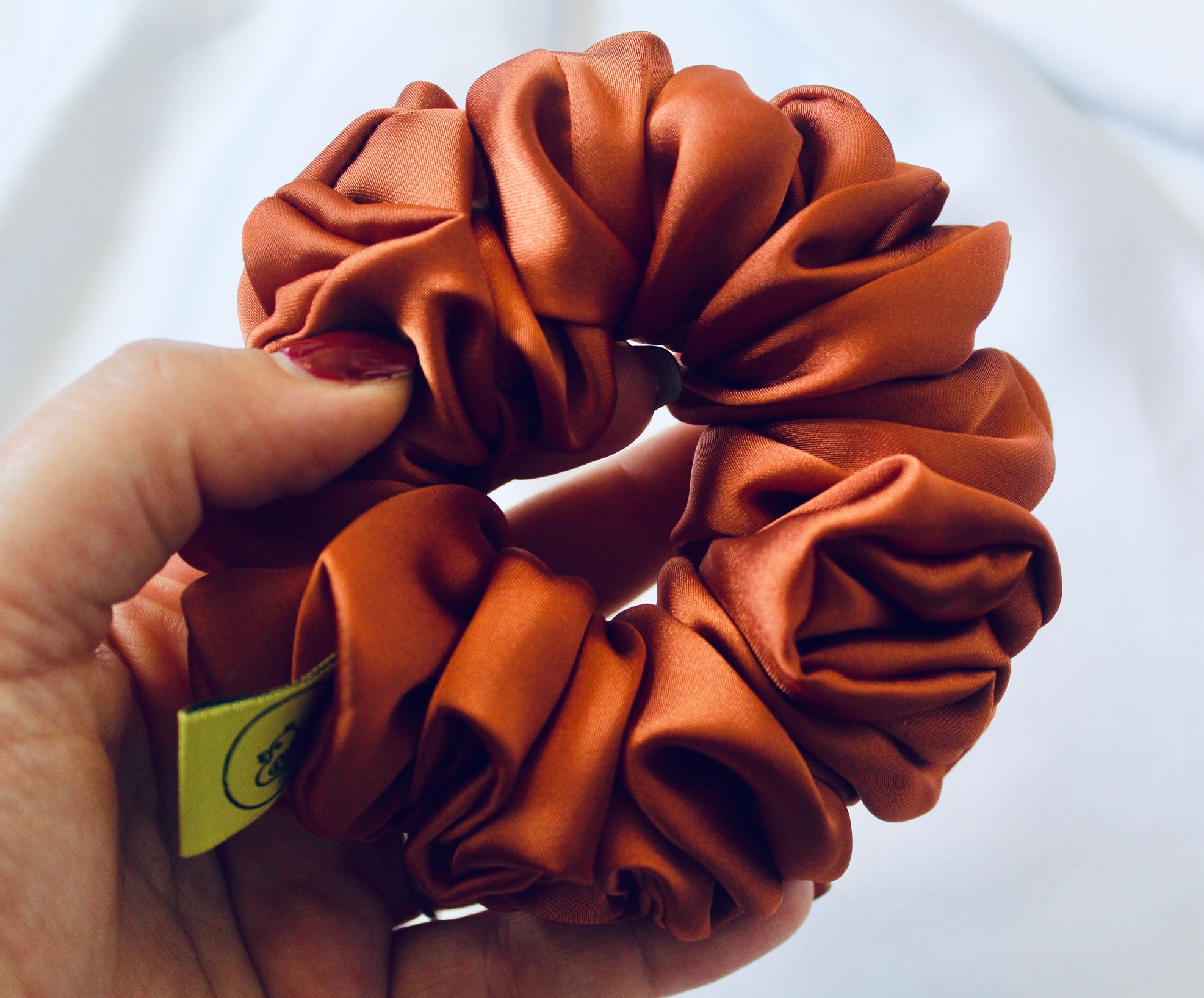 A satin scrunchie in a warm burnt orange shade with a smooth glossy texture and a ruched design.  This scrunchie is made from satin giving it a silky and lightweight feel.  The rich burnt orange colour captures the warmth of autumn leaves, making it perfect seasonal accessory.  The gathered fabric sits on an elastic core, creating a full ruffled look while keeping your hairstyle secure and comfortable.