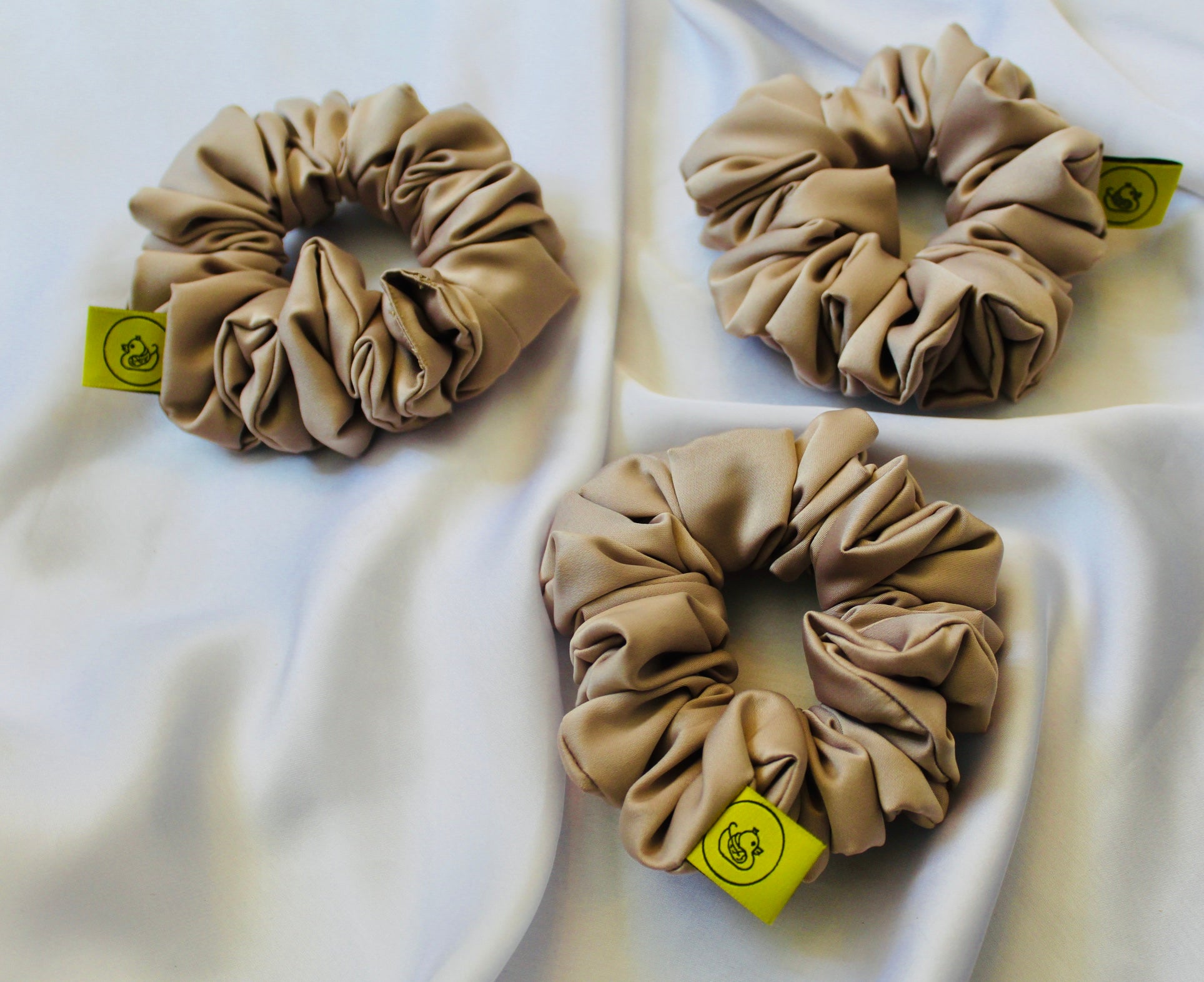 A satin hair scrunchie in a soft taupe beigge with a smooth glossy texture and a ruched design.  This scrunchie is made from satin, giving it a silky, luxurious feel.  The soft taupe shade is a neutral tone that pairs well with any outfit, making it a timeless everyday essential.  It's fabric is gathered along an elastic core, creating a full, ruffled look while staying secure and comfortable in the hair.