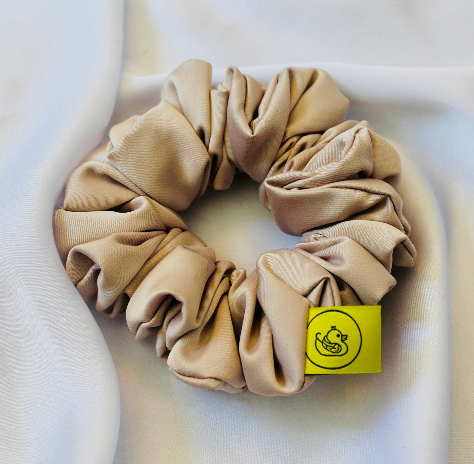 A satin hair scrunchie in a soft taupe beigge with a smooth glossy texture and a ruched design.  This scrunchie is made from satin, giving it a silky, luxurious feel.  The soft taupe shade is a neutral tone that pairs well with any outfit, making it a timeless everyday essential.  It's fabric is gathered along an elastic core, creating a full, ruffled look while staying secure and comfortable in the hair.