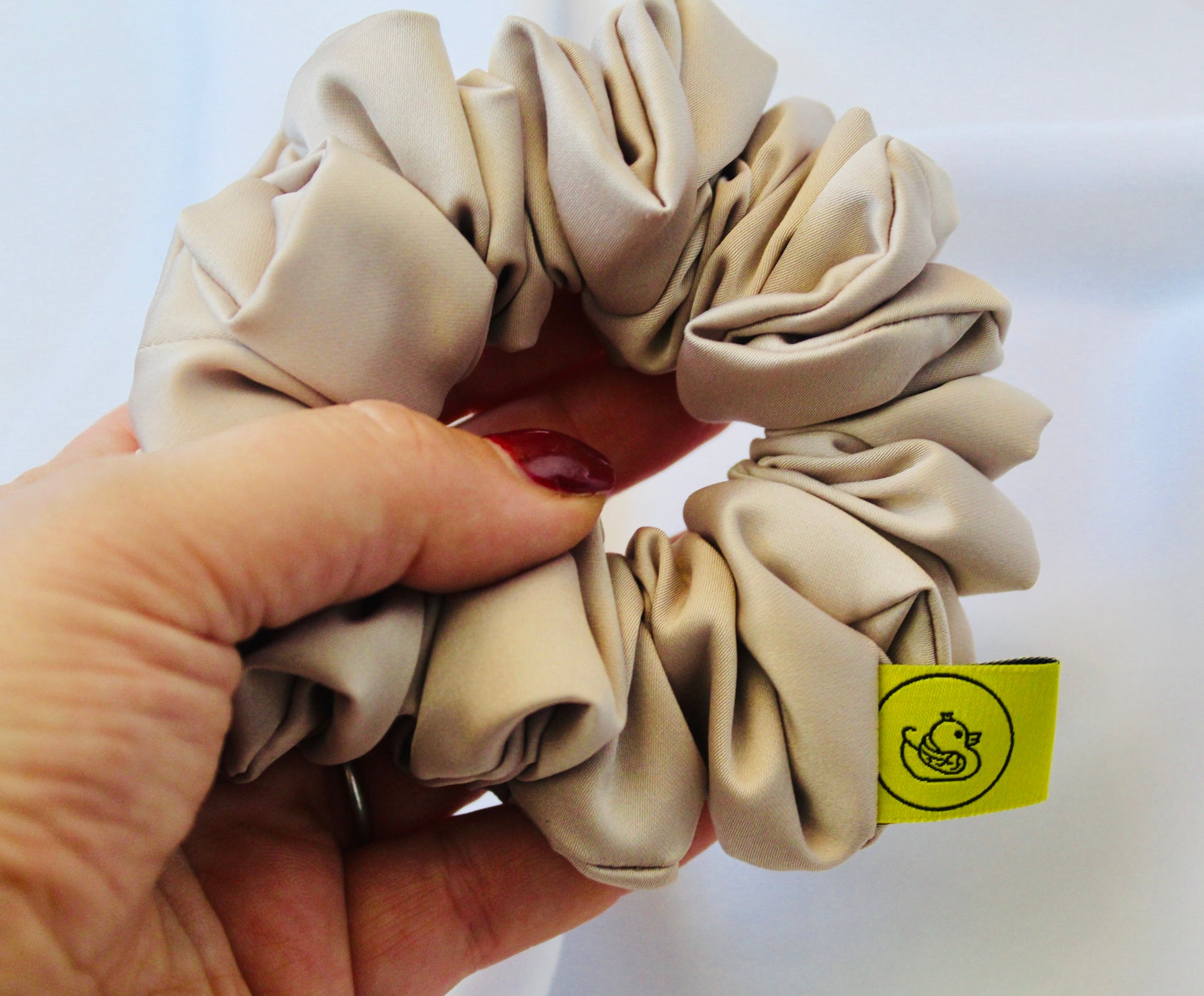 A satin hair scrunchie in a soft taupe beigge with a smooth glossy texture and a ruched design.  This scrunchie is made from satin, giving it a silky, luxurious feel.  The soft taupe shade is a neutral tone that pairs well with any outfit, making it a timeless everyday essential.  It's fabric is gathered along an elastic core, creating a full, ruffled look while staying secure and comfortable in the hair.