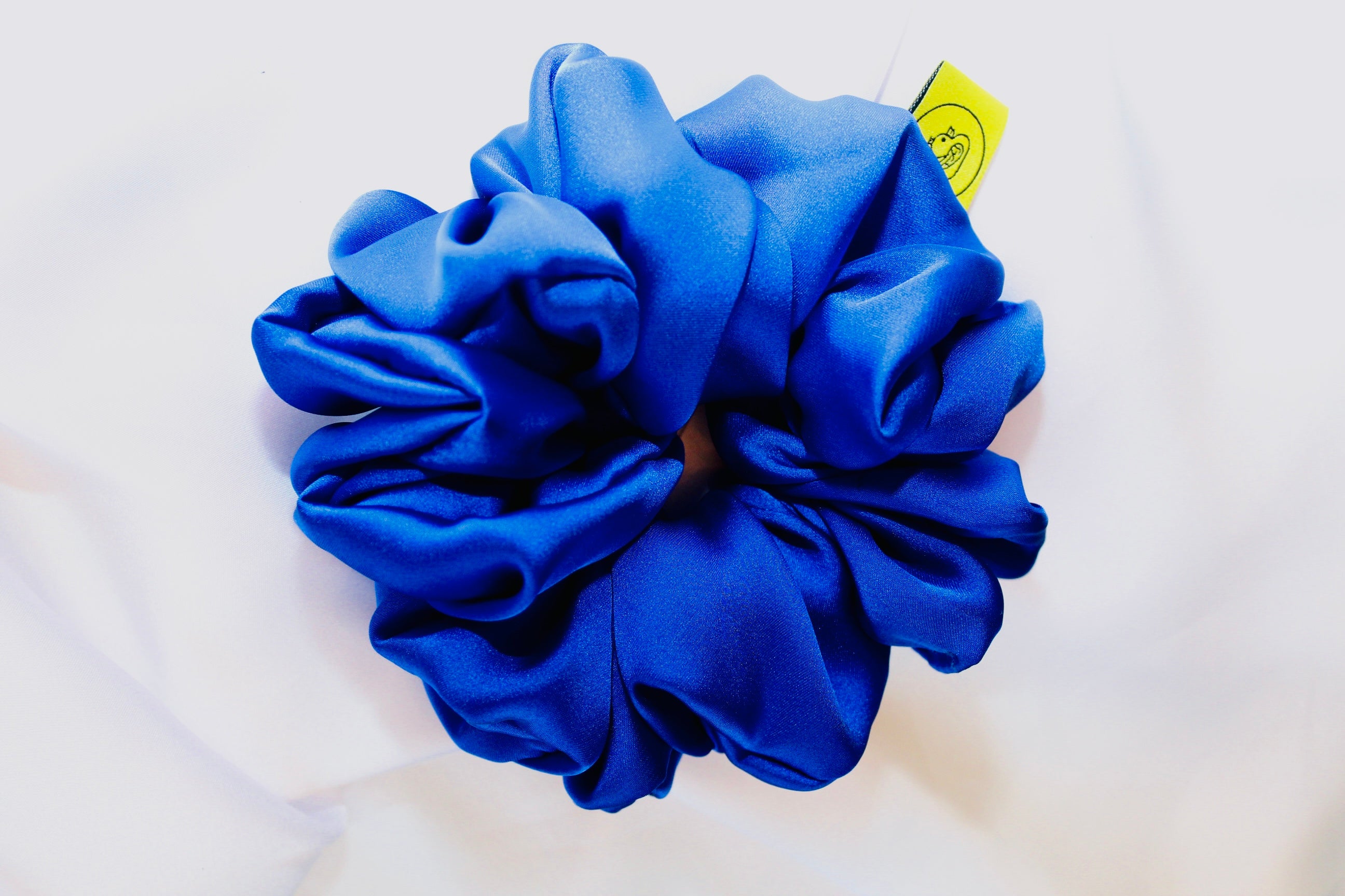 This shows a luxurious scrunchie made of silky textured fabric in a royal blue giving it an elegant and slightly reflective appearance.The scrunchie is gathered and voluminous creating soft folds and a plush look.A small yellow label tag with a picture of a duck is attached to the scrunchie adding a touch of branding.The overall aesthetic is soft, elegant and stylish. Perfect for those with sensitive scalp or hair loss