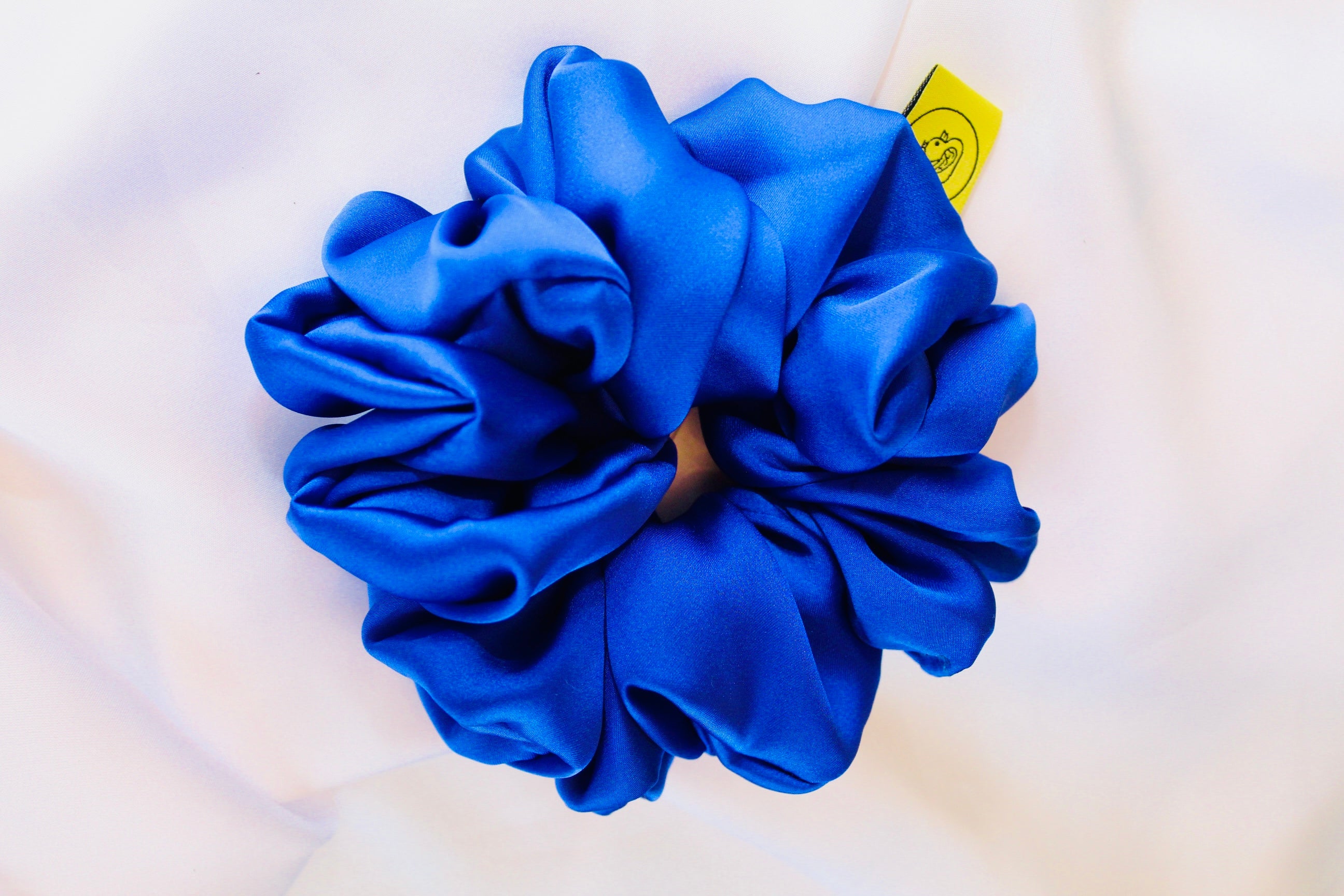 This shows a luxurious scrunchie made of silky textured fabric in a royal blue giving it an elegant and slightly reflective appearance.The scrunchie is gathered and voluminous creating soft folds and a plush look.A small yellow label tag with a picture of a duck is attached to the scrunchie adding a touch of branding.The overall aesthetic is soft, elegant and stylish. Perfect for those with sensitive scalp or hair loss
