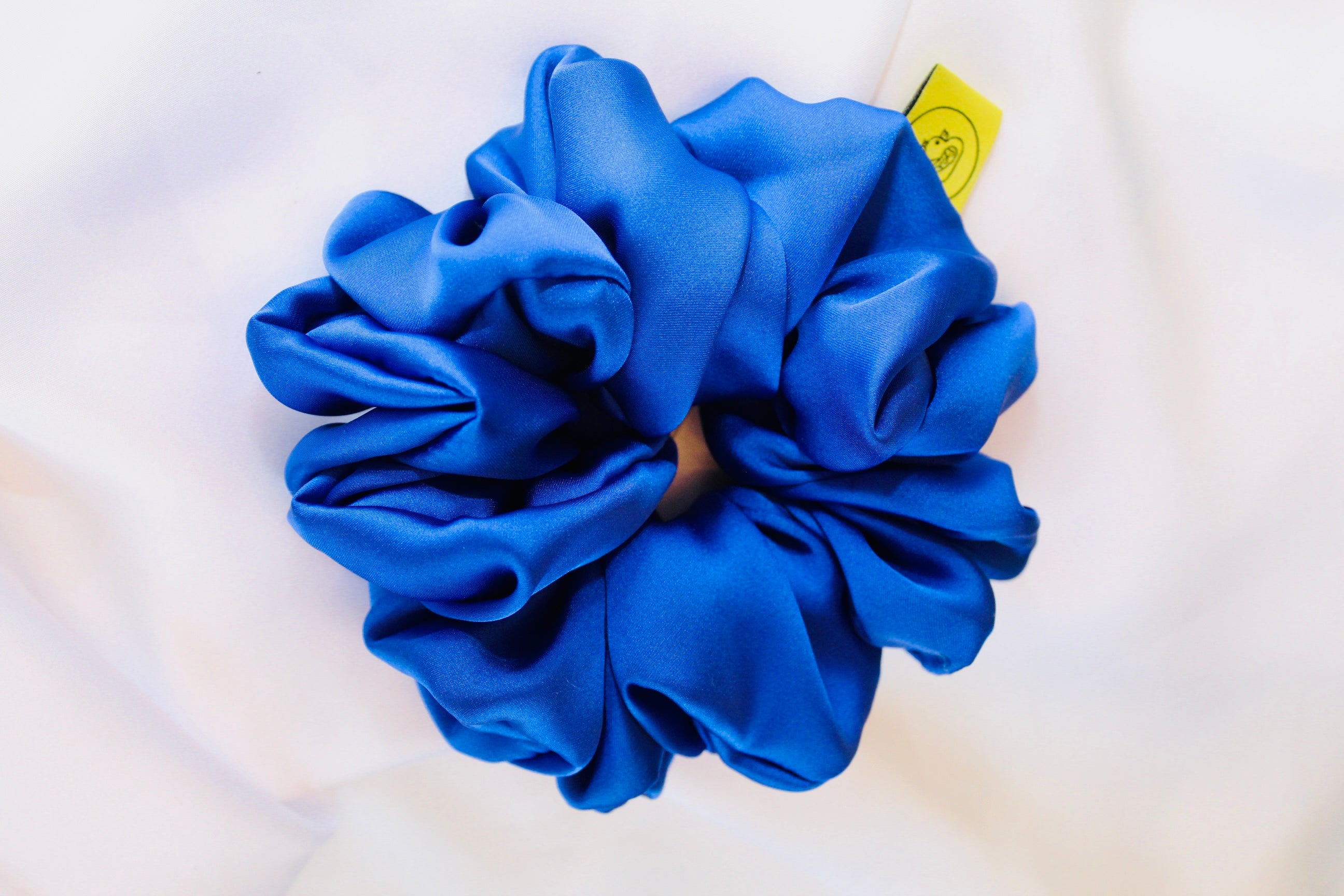 This shows a luxurious scrunchie made of silky textured fabric in a royal blue giving it an elegant and slightly reflective appearance.The scrunchie is gathered and voluminous creating soft folds and a plush look.A small yellow label tag with a picture of a duck is attached to the scrunchie adding a touch of branding.The overall aesthetic is soft, elegant and stylish. Perfect for those with sensitive scalp or hair loss