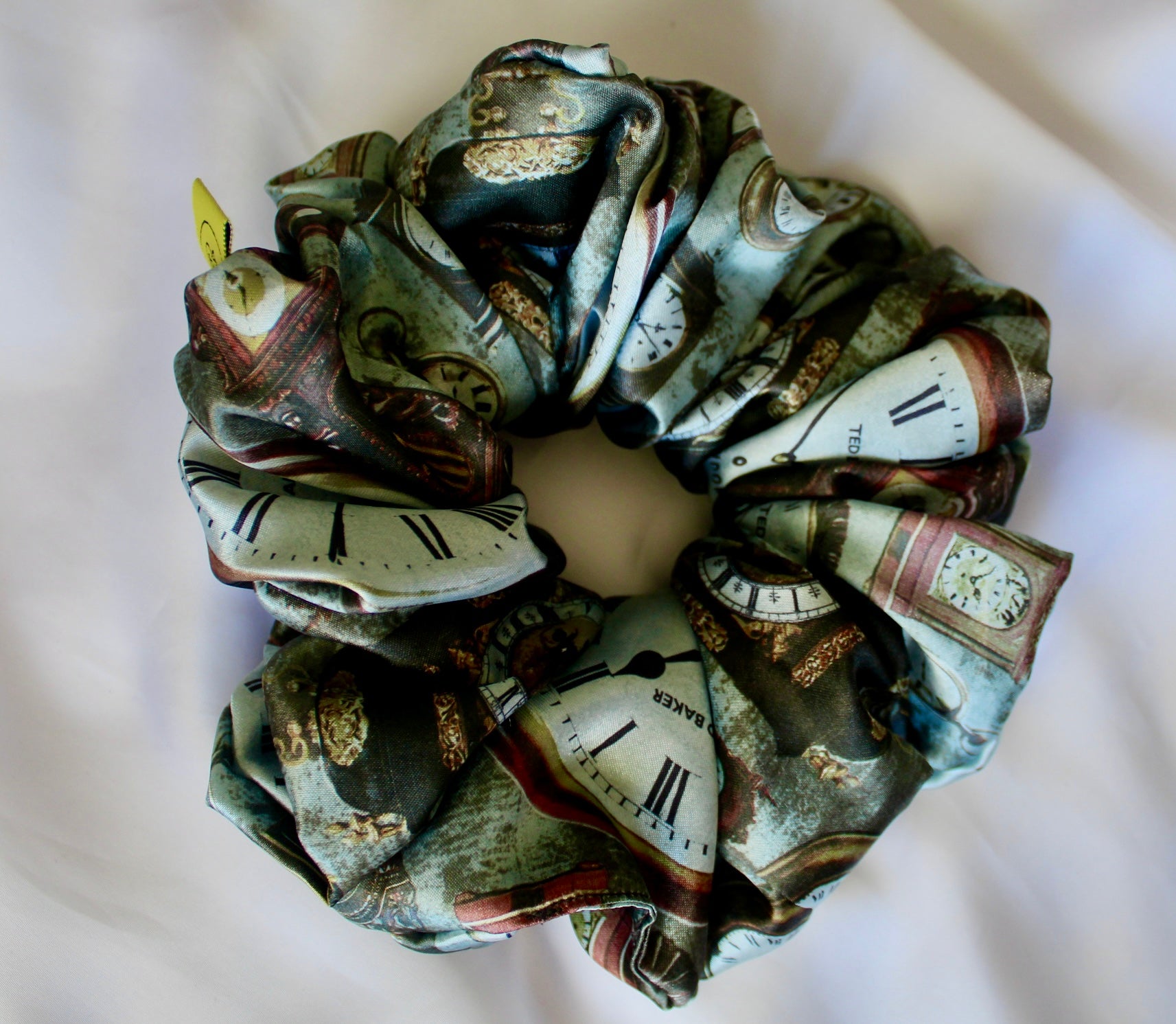 This image features a handmade regular sized hair scrunchie with a vintage clock print on a satin fabric.  The scrunchie is designed with a mix of antique- style timepieces, numbers and text elements, giving it a steampunk and Victorian Aesthetic.  The primary colours include shades of brown and beige. The fabric has a slight sheen adding a luxurious touch to the accessory. Ideal for those looking for a statement hair accessory.