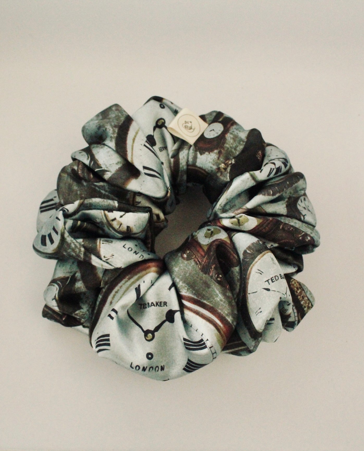 This image features a handmade regular sized hair scrunchie with a vintage clock print on a satin fabric.  The scrunchie is designed with a mix of antique- style timepieces, numbers and text elements, giving it a steampunk and Victorian Aesthetic.  The primary colours include shades of brown and beige. The fabric has a slight sheen adding a luxurious touch to the accessory. Ideal for those looking for a statement hair accessory.