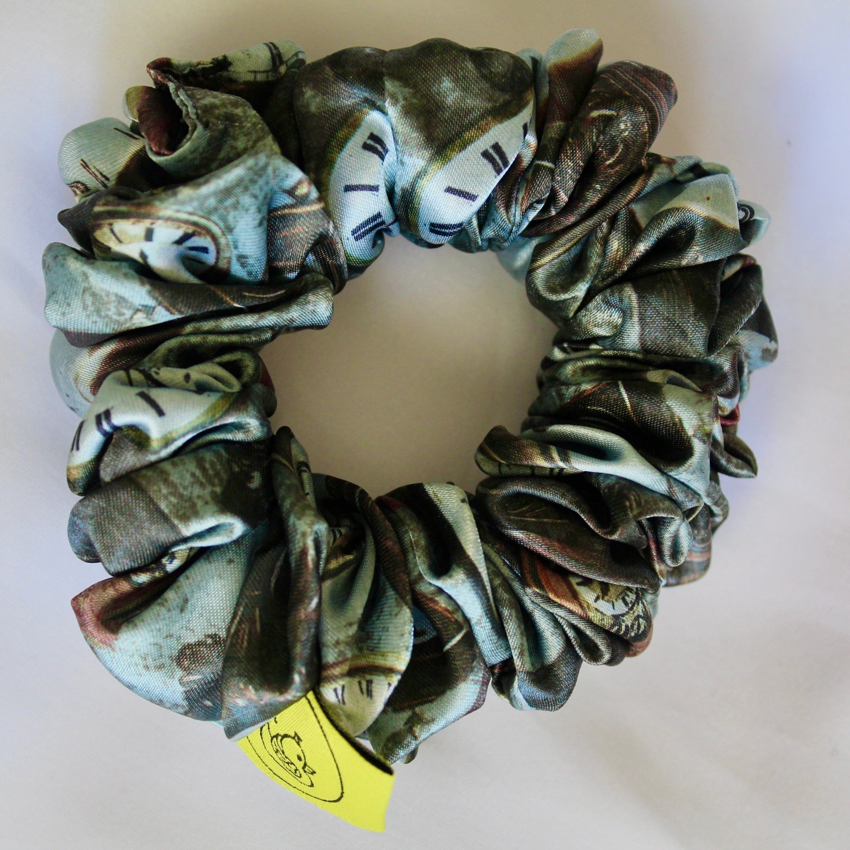 This image features a handmade regular sized hair scrunchie with a vintage clock print on a satin fabric.  The scrunchie is designed with a mix of antique- style timepieces, numbers and text elements, giving it a steampunk and Victorian Aesthetic.  The primary colours include shades of brown and beige. The fabric has a slight sheen adding a luxurious touch to the accessory. Ideal for those looking for a statement hair accessory.