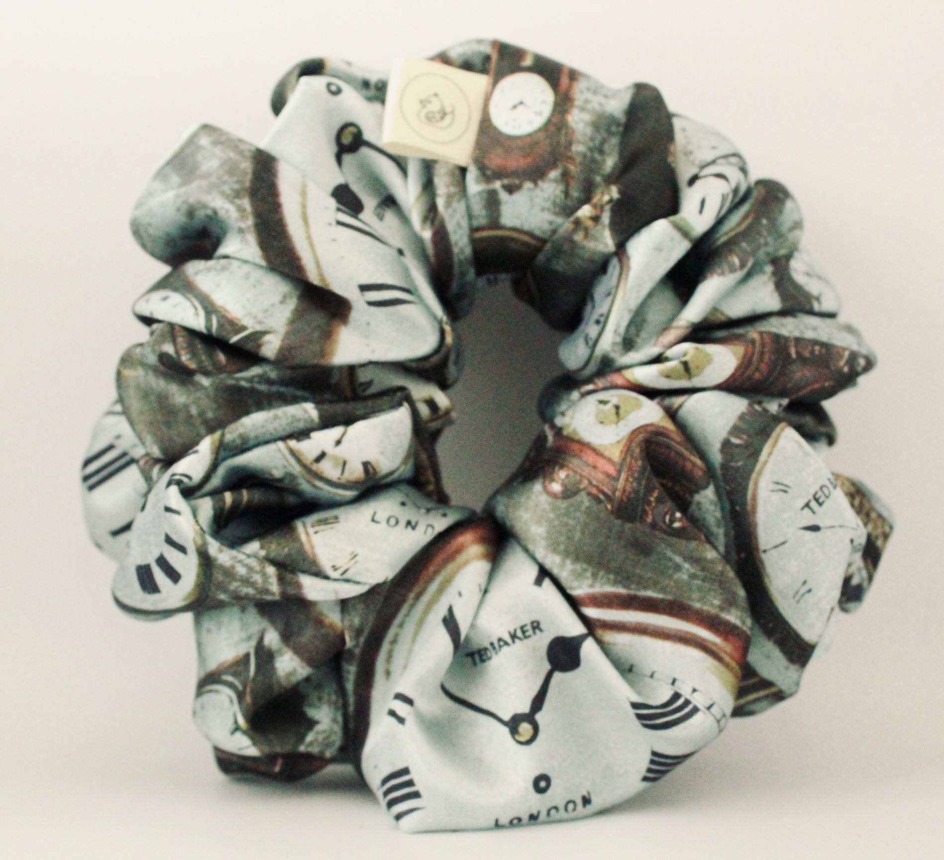 This image features a handmade regular sized hair scrunchie with a vintage clock print on a satin fabric.  The scrunchie is designed with a mix of antique- style timepieces, numbers and text elements, giving it a steampunk and Victorian Aesthetic.  The primary colours include shades of brown and beige. The fabric has a slight sheen adding a luxurious touch to the accessory. Ideal for those looking for a statement hair accessory.