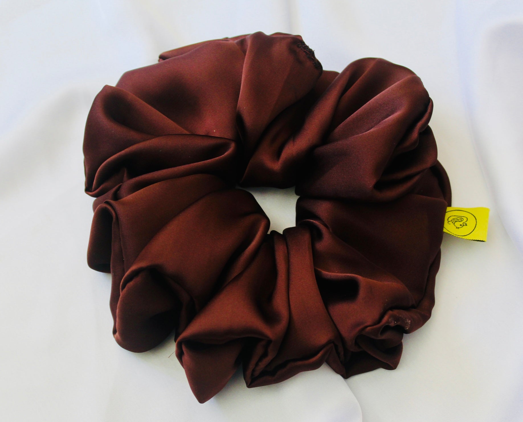A satin scrunchie in a rich chocolate brown shade with a smooth and shiny texture and a ruched design.  This scrunchie is made from glossy satin giving it a soft, luxurious touch.  The deep chocolate brown colour is both bold and neutral, making it easy to style with a wide range of outfits.  The fabric is softly ruched over elastic, creating volume while remaining comfortable to wear.