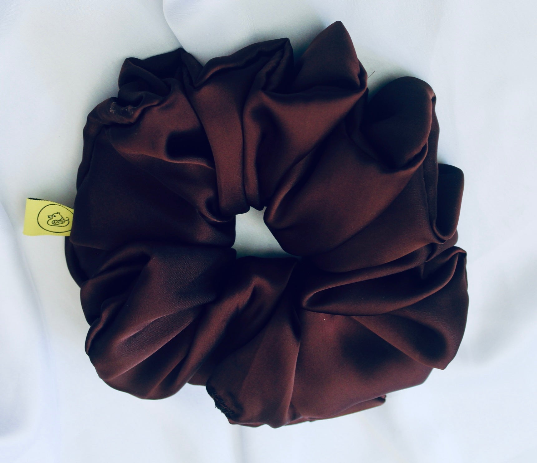 A satin scrunchie in a rich chocolate brown shade with a smooth and shiny texture and a ruched design.  This scrunchie is made from glossy satin giving it a soft, luxurious touch.  The deep chocolate brown colour is both bold and neutral, making it easy to style with a wide range of outfits.  The fabric is softly ruched over elastic, creating volume while remaining comfortable to wear.