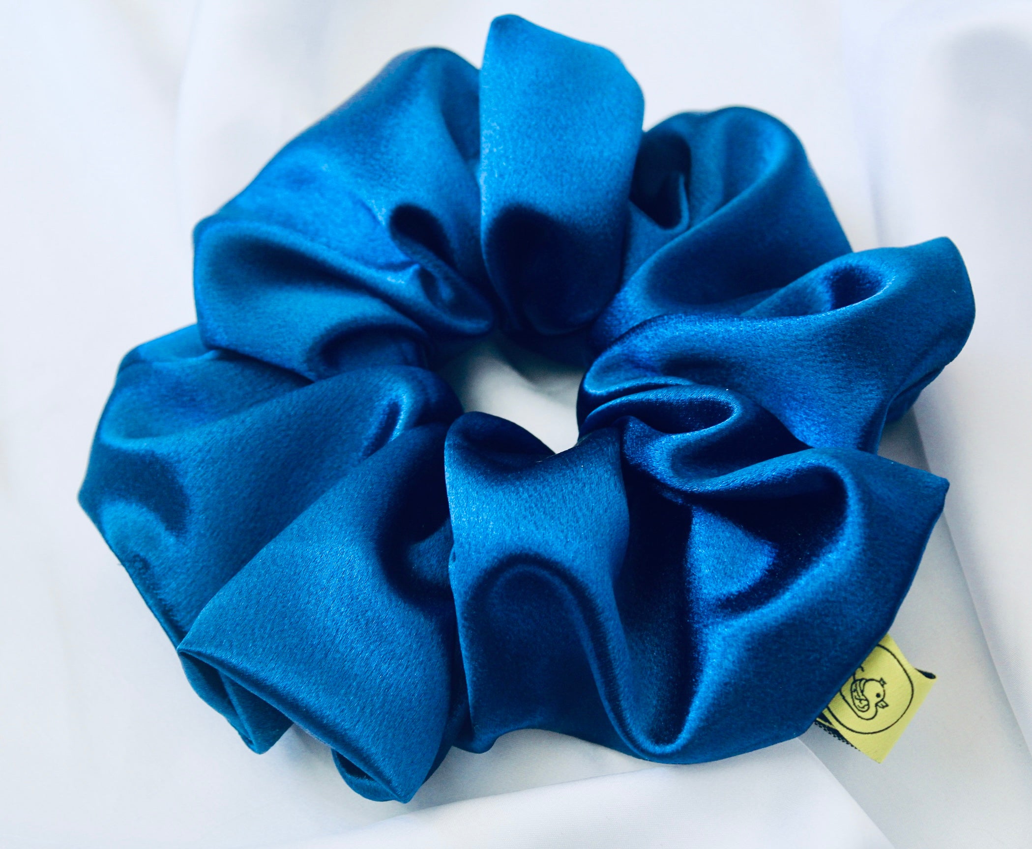 A satin scrunchie in a striking royal blue shade with a silky glossy texture and a ruched design.  Crafted from satin this scrunchie feels smooth and cool to the touch.  The bold royal blue colour stands out beautifully, making it a statement piece for both casual and evening looks.  The gathered satin fabric creates a full textured effect over the elastic ensuring a secure hold.