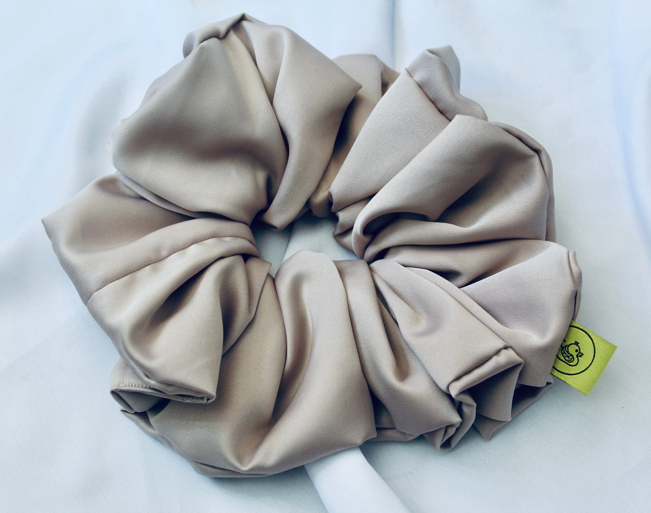 A satin hair scrunchie in a soft taupe beigge with a smooth glossy texture and a ruched design.  This scrunchie is made from satin, giving it a silky, luxurious feel.  The soft taupe shade is a neutral tone that pairs well with any outfit, making it a timeless everyday essential.  It's fabric is gathered along an elastic core, creating a full, ruffled look while staying secure and comfortable in the hair.