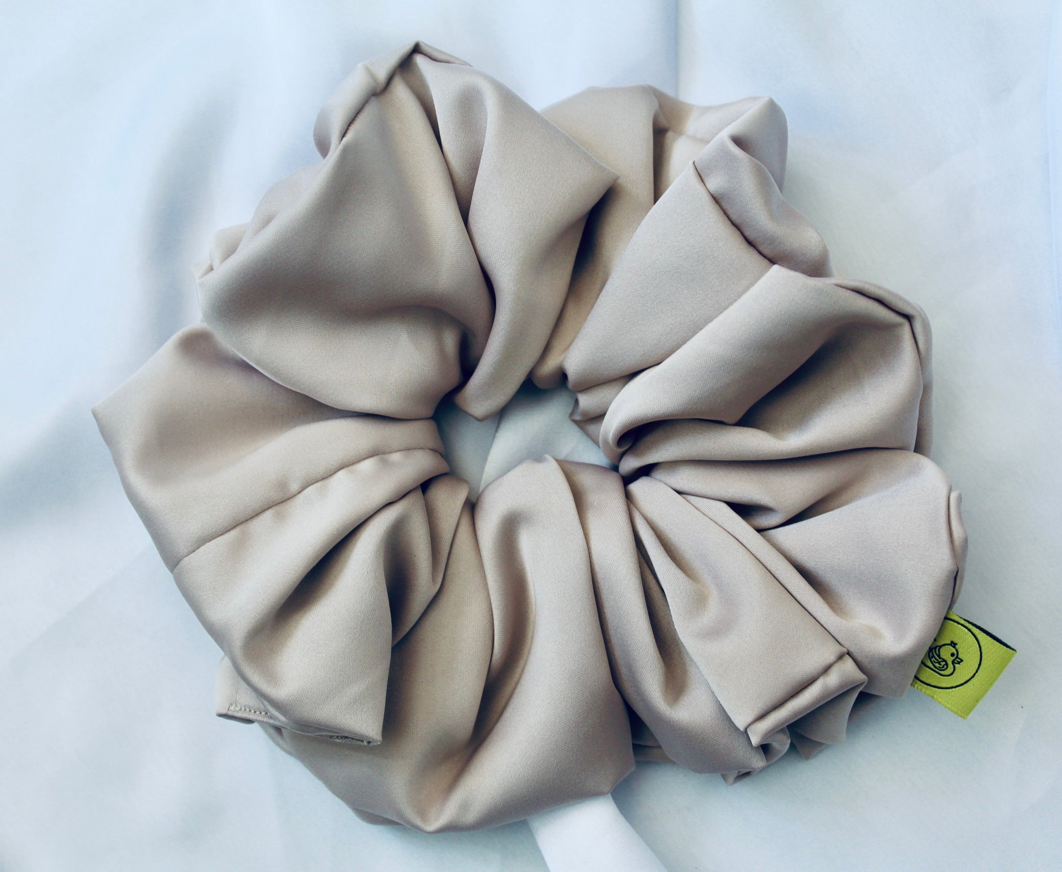 A satin hair scrunchie in a soft taupe beigge with a smooth glossy texture and a ruched design.  This scrunchie is made from satin, giving it a silky, luxurious feel.  The soft taupe shade is a neutral tone that pairs well with any outfit, making it a timeless everyday essential.  It's fabric is gathered along an elastic core, creating a full, ruffled look while staying secure and comfortable in the hair.