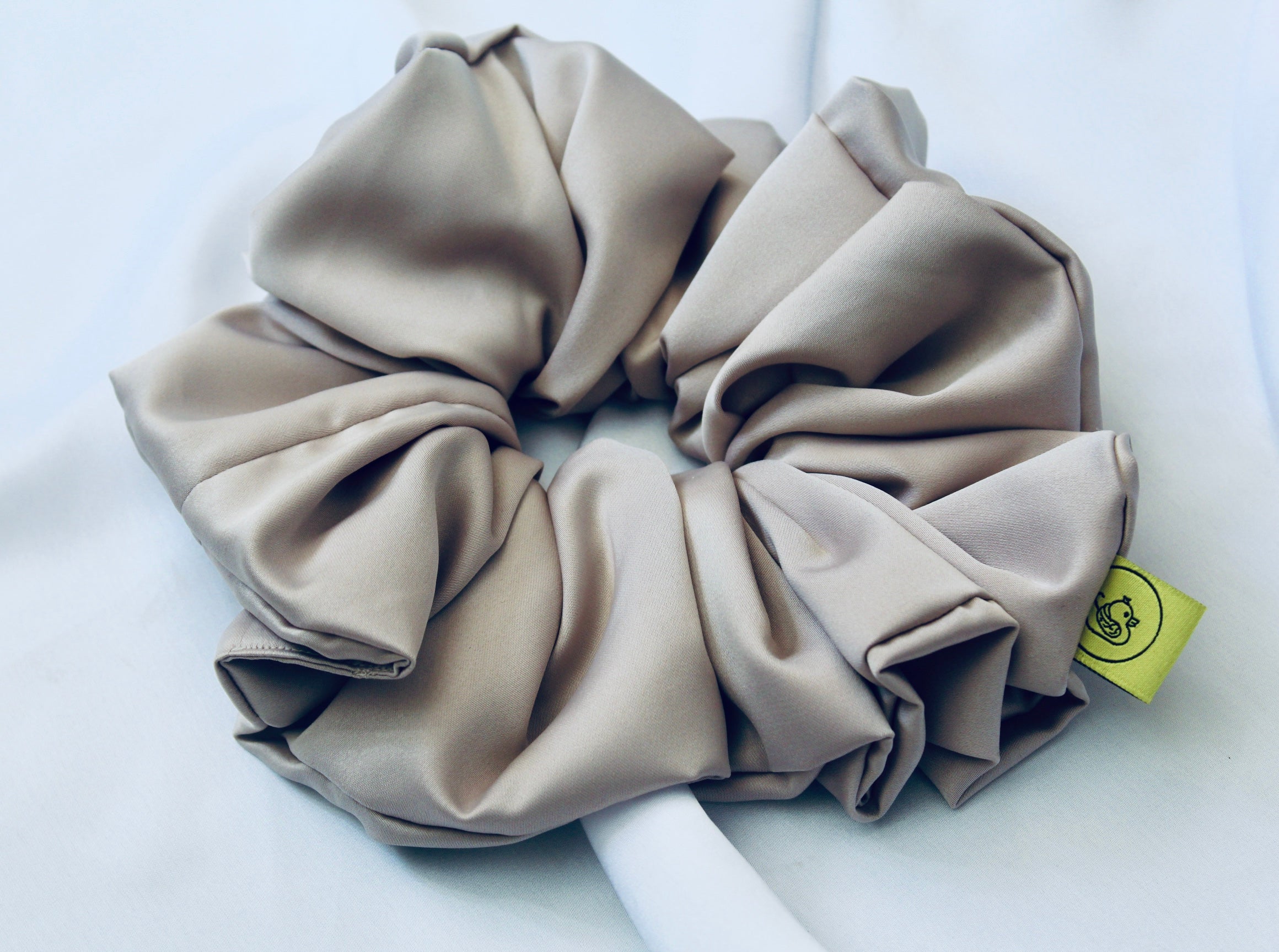A satin hair scrunchie in a soft taupe beigge with a smooth glossy texture and a ruched design.  This scrunchie is made from satin, giving it a silky, luxurious feel.  The soft taupe shade is a neutral tone that pairs well with any outfit, making it a timeless everyday essential.  It's fabric is gathered along an elastic core, creating a full, ruffled look while staying secure and comfortable in the hair.