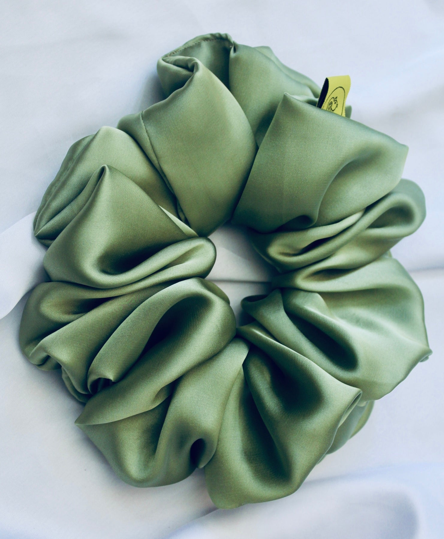 A satin scrunchie in a light green shade with a silky, glossy finish and a ruched design.  Crafted from smooth satin, this scrunchie feels soft and sleek against the hair.  The green tone has a timeless, earthy quality, perfect for both everyday wear and festive styling.  Its ruched fabric is stretched over an elastic band, providing a secure yet comfortable fit.