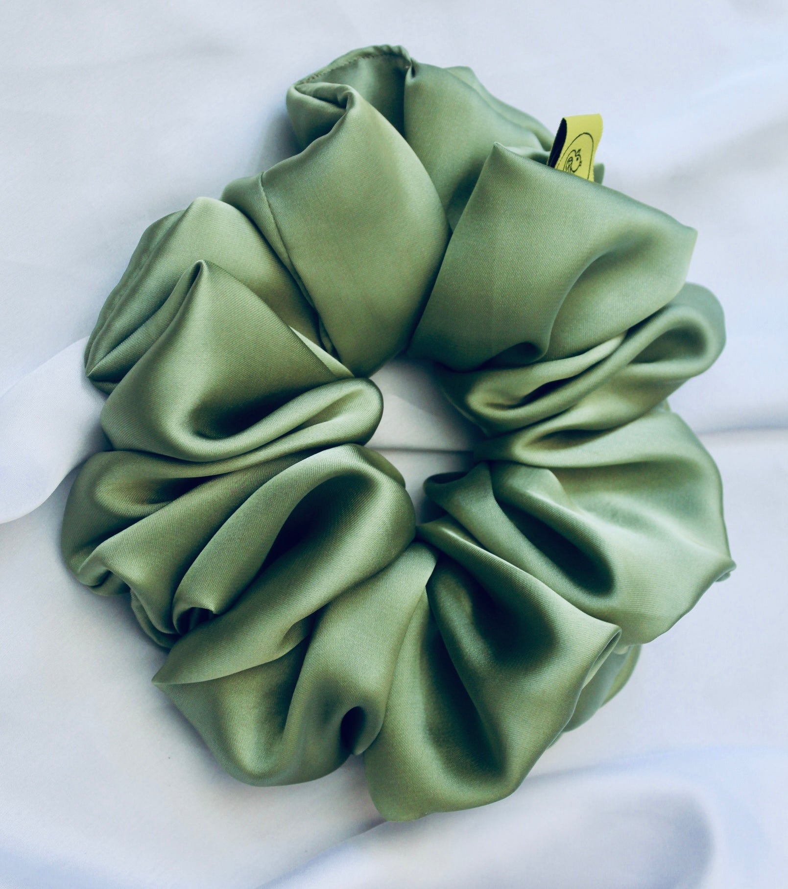 A satin scrunchie in a light green shade with a silky, glossy finish and a ruched design.  Crafted from smooth satin, this scrunchie feels soft and sleek against the hair.  The green tone has a timeless, earthy quality, perfect for both everyday wear and festive styling.  Its ruched fabric is stretched over an elastic band, providing a secure yet comfortable fit.