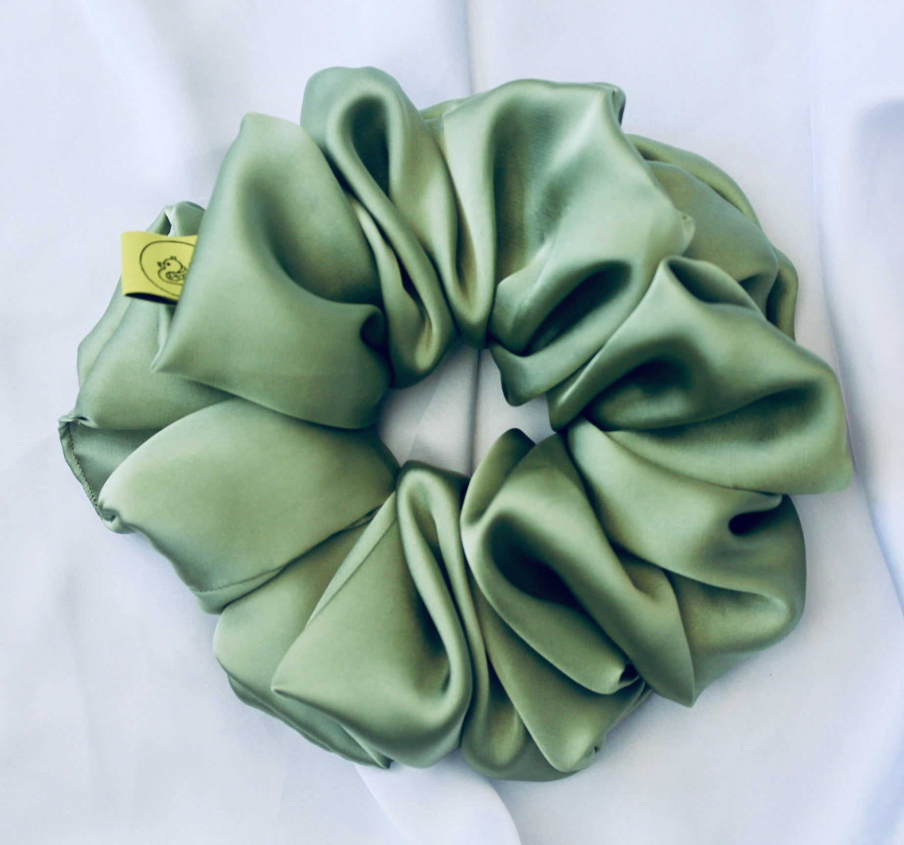 A satin scrunchie in a light green shade with a silky, glossy finish and a ruched design.  Crafted from smooth satin, this scrunchie feels soft and sleek against the hair.  The green tone has a timeless, earthy quality, perfect for both everyday wear and festive styling.  Its ruched fabric is stretched over an elastic band, providing a secure yet comfortable fit.